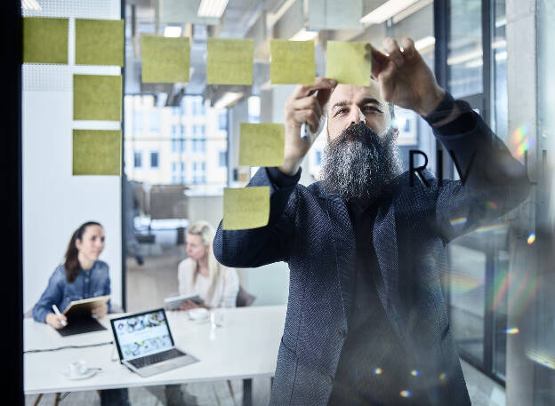 Unternehmensfoto Mann mit grauem Anzug klebt Post it an Glaswand Mitarbeiterinnen im Hintergrund Employer Branding - Businessfotograf Businessfotografie Christian Vorhofer