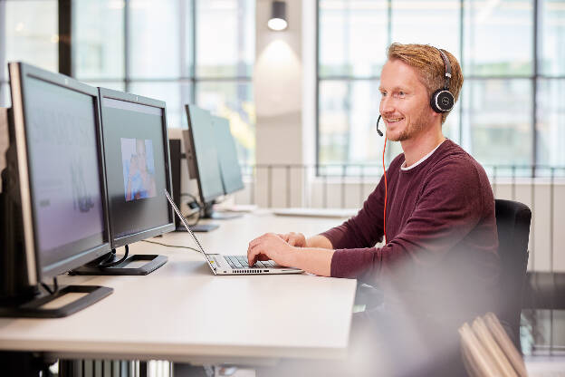 Mann sitzt mit Headset, Laptop und mehreren Bildschirmen am Tisch, modernes Gebäude - Businessfotograf Businessfotografie Christian Vorhofer