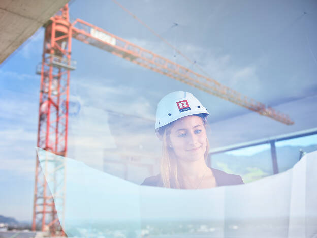 Unternehmensfoto Architektin mit Helm und Bauplan hinter einem Fenster in der Scheibe spiegelt sich ein Baukran - Businessfotograf Businessfotografie Christian Vorhofer